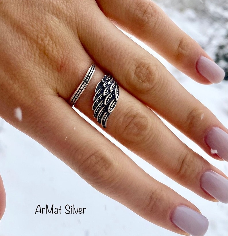 May include: Two silver rings on a finger. The ring on the index finger is a simple band. The ring on the middle finger is a wing design.