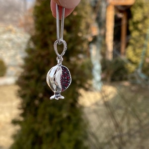 May include: A silver chain necklace with a pomegranate pendant. The pendant is made of silver and has red gemstones that resemble pomegranate seeds.