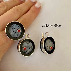 Op de afbeelding: Een zilveren ring en oorbellenset met een zwart-wit patroon en een rode steentjesaccent. De tekst "ArMat Silver" is zichtbaar in de afbeelding.