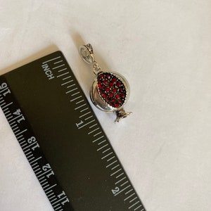 May include: A silver charm in the shape of a pomegranate with red crystal accents. The charm is approximately 2.5 cm long and is shown next to a ruler.