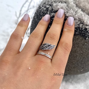 May include: A silver ring with a wing design. The ring is on a person's finger. The background is snowy.