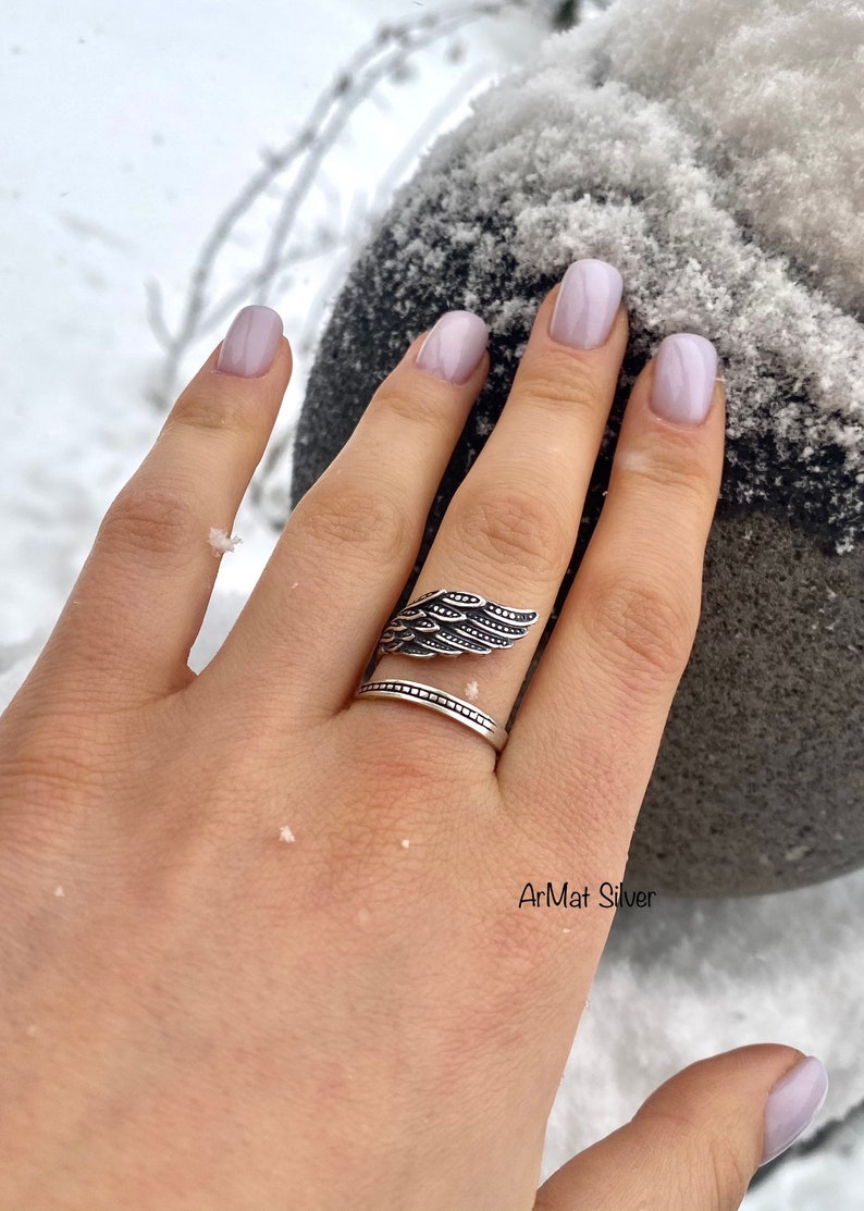 May include: A silver ring with a wing design. The ring is on a person's finger. The background is snowy.