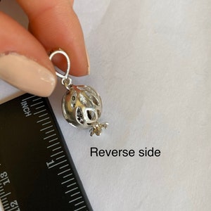May include: Silver charm in the shape of a pomegranate with a detailed design. The charm is approximately 2.5 cm in diameter and has a loop for attaching to a bracelet or necklace. The back of the charm is shown in the image. Reverse side.