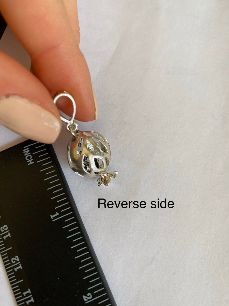 May include: Silver charm in the shape of a pomegranate with a detailed design. The charm is approximately 2.5 cm in diameter and has a loop for attaching to a bracelet or necklace. The back of the charm is shown in the image. Reverse side.