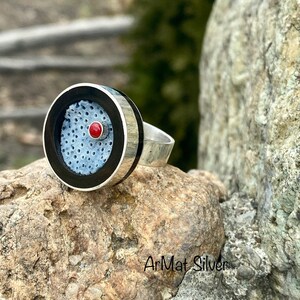 Op de afbeelding: Een zilveren ring met een zwart-wit cirkelvormig ontwerp met een rode edelsteen. De ring ligt op een grijze steen.