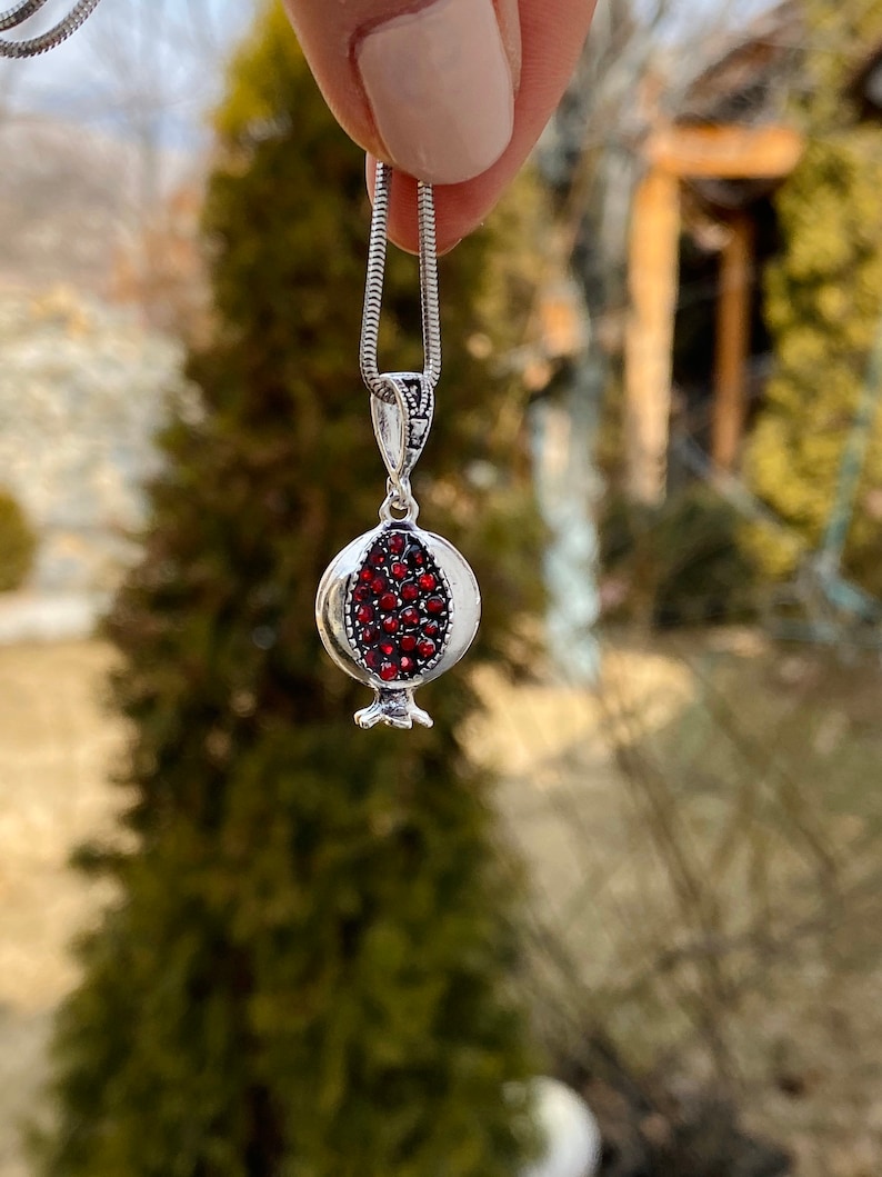 May include: A silver chain necklace with a pomegranate pendant. The pendant is made of silver and has red gemstones that resemble pomegranate seeds.