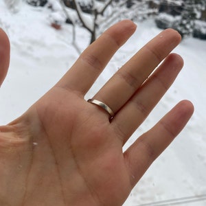 May include: A close-up of a hand wearing a simple silver band ring. The ring is on the ring finger of the left hand. The hand is against a blurry background of a snowy landscape.