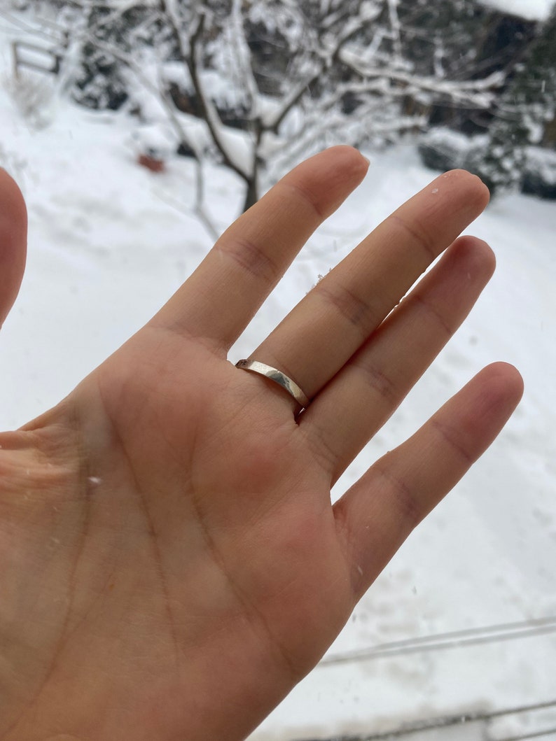 May include: A close-up of a hand wearing a simple silver band ring. The ring is on the ring finger of the left hand. The hand is against a blurry background of a snowy landscape.