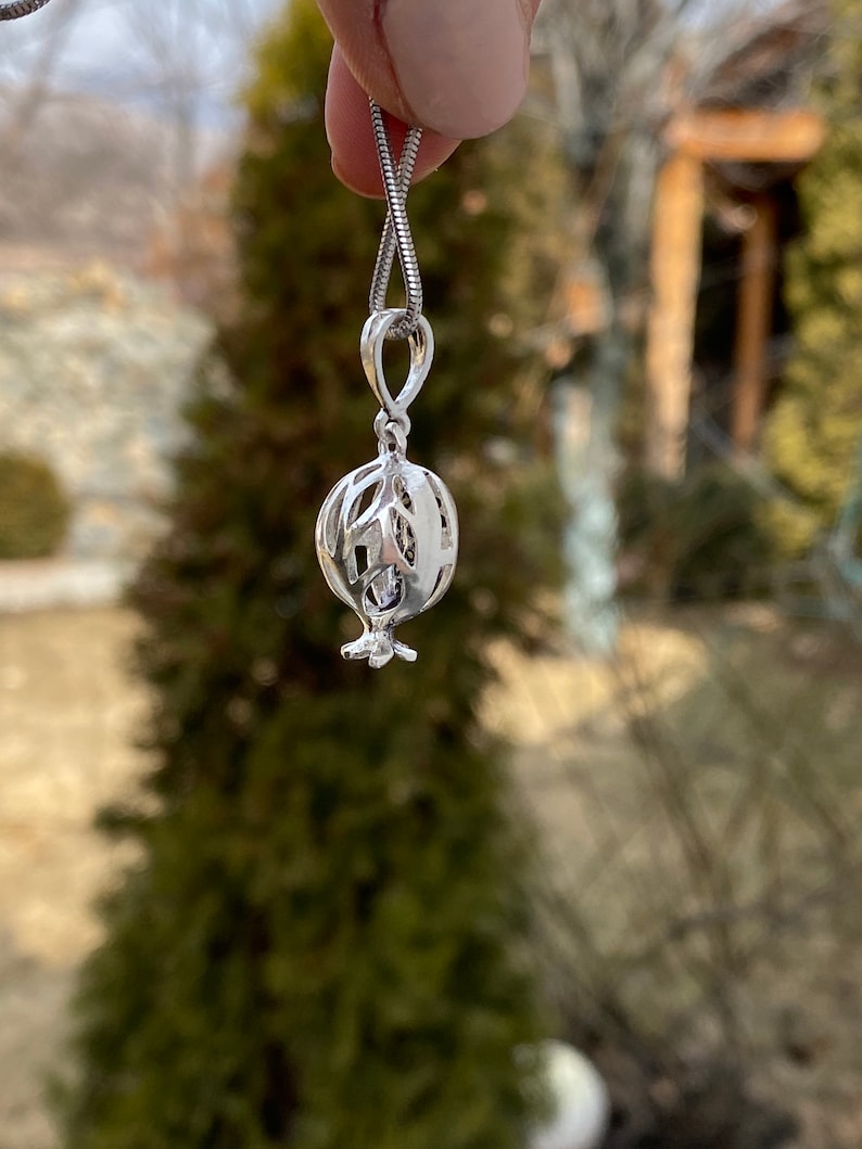 May include: A silver necklace featuring a delicate, openwork pomegranate pendant. The pendant is detailed with a small, clear gemstone.