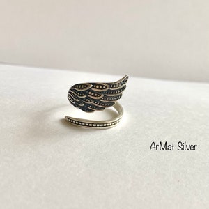 May include: A silver ring with a detailed wing design. The ring is open and wraps around the finger. The text "ArMat Silver" is visible below the ring.