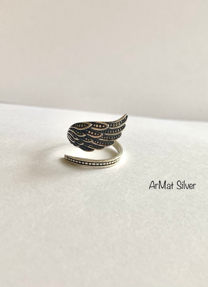 May include: A silver ring with a detailed wing design. The ring is open and wraps around the finger. The text "ArMat Silver" is visible below the ring.