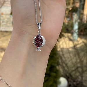 May include: A silver chain necklace with a pomegranate pendant. The pendant is made of silver and has red gemstones that resemble pomegranate seeds.