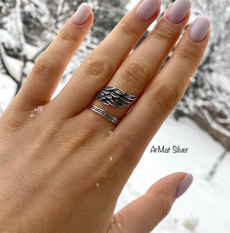 May include: A silver ring with an angel wing design. The ring is on a finger with a plain silver band underneath. The text "ArMat Silver" is visible in the lower right corner of the image.