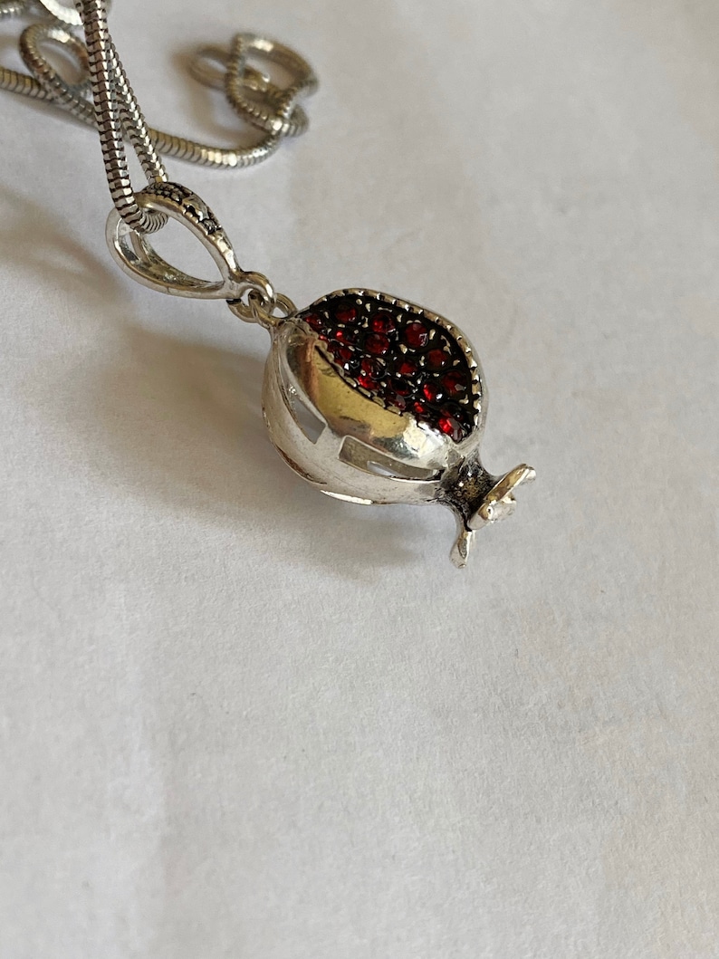 May include: A silver necklace with a pomegranate pendant. The pendant is decorated with red gemstones.