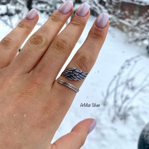 May include: A silver ring with a wing design. The ring is on a finger with light pink nail polish. The background is blurry and shows snow.
