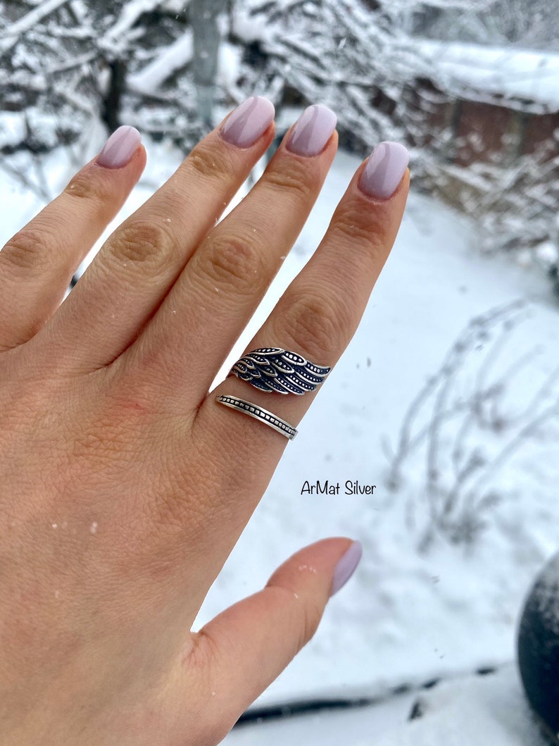 May include: A silver ring with a wing design. The ring is on a finger with light pink nail polish. The background is blurry and shows snow.