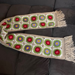 May include: A crocheted scarf with a fringe on both ends. The scarf is made of beige yarn and features a repeating pattern of colorful flowers in shades of red, green, purple, and white.