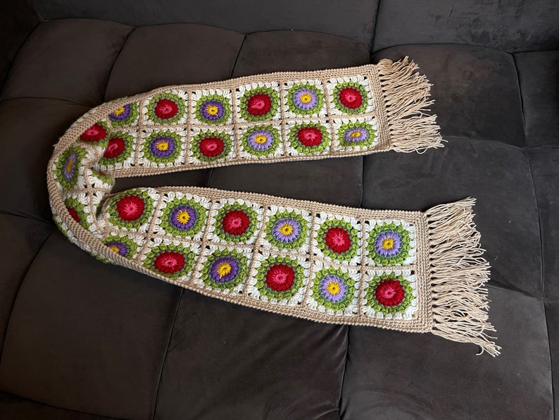 May include: A crocheted scarf with a fringe on both ends. The scarf is made of beige yarn and features a repeating pattern of colorful flowers in shades of red, green, purple, and white.