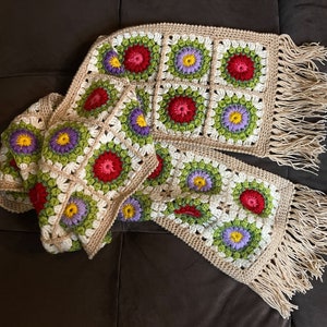 May include: A crocheted scarf with a fringe, featuring a pattern of colorful flowers in shades of red, green, purple, and yellow.