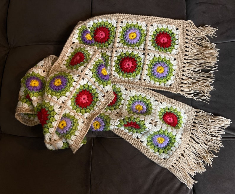 May include: A crocheted scarf with a fringe, featuring a pattern of colorful flowers in shades of red, green, purple, and yellow.