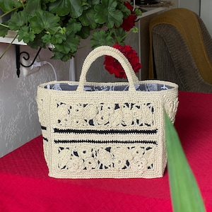 May include: A beige crocheted tote bag with floral patterns and black stripes. The bag has a rectangular shape with a handle and a patterned interior. The bag is sitting on a red surface.