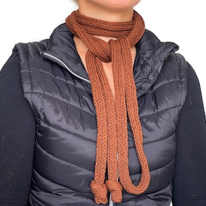 May include: A brown knitted scarf with a long, loose knot. The scarf is worn over a black quilted vest.