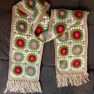 May include: A crocheted scarf with a floral pattern in shades of red, green, purple, and yellow. The scarf has a fringe on both ends.