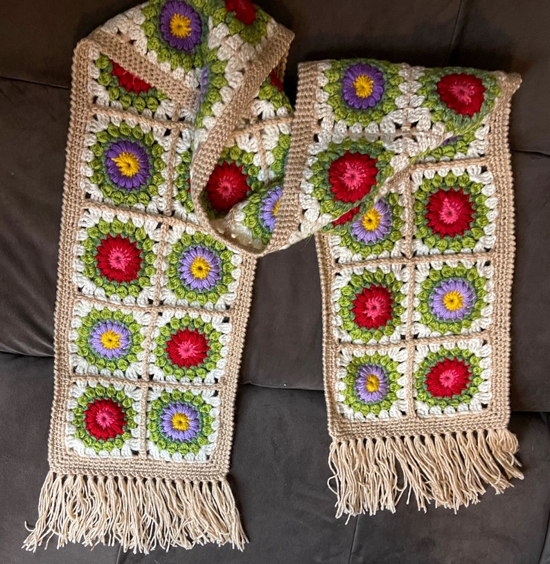 May include: A crocheted scarf with a floral pattern in shades of red, green, purple, and yellow. The scarf has a fringe on both ends.