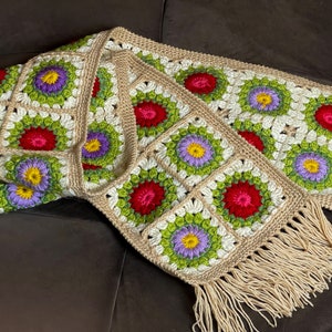 May include: A crocheted throw blanket with a fringe border. The blanket is made of beige yarn and features a repeating pattern of colorful flowers.