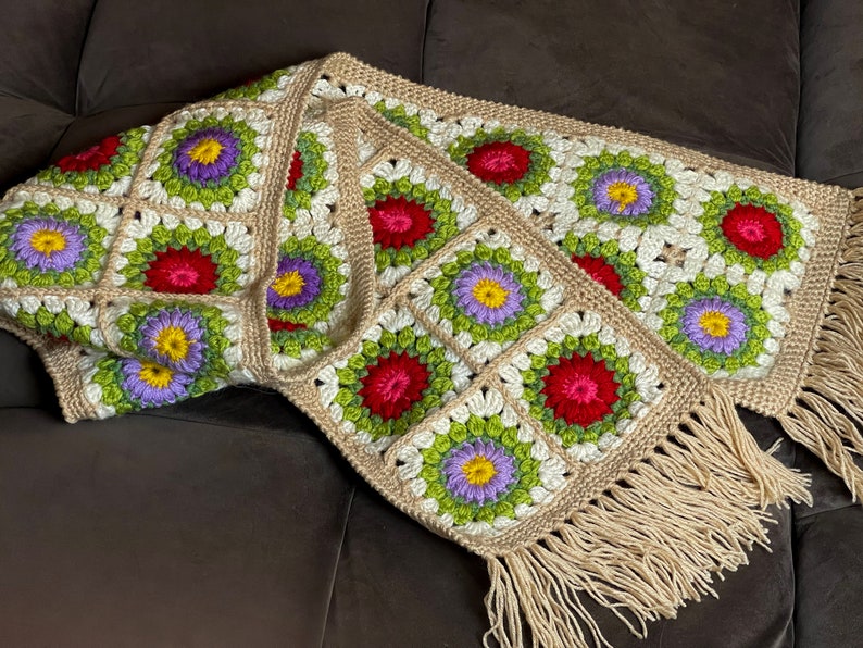 May include: A crocheted throw blanket with a fringe border. The blanket is made of beige yarn and features a repeating pattern of colorful flowers.