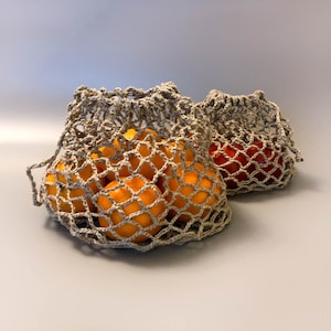 Könnte beinhalten: Zwei beigefarbene Häkelnetztaschen, eine gefüllt mit orangenen Zitrusfrüchten und die andere mit einer roten Tomate.