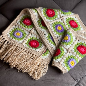 May include: A crocheted scarf with a fringe border. The scarf is made of beige yarn and features a repeating pattern of colorful flowers.