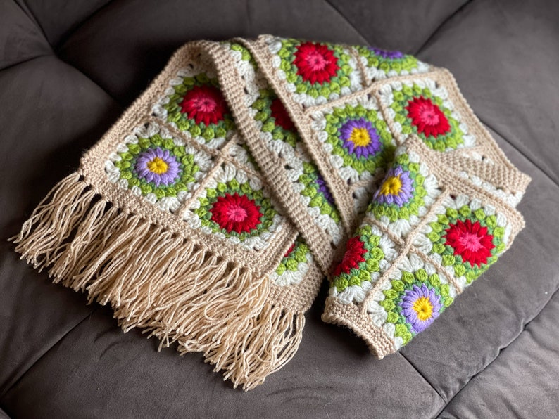 May include: A crocheted scarf with a fringe border. The scarf is made of beige yarn and features a repeating pattern of colorful flowers.