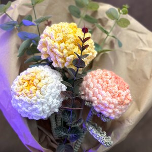 May include: A bouquet of crocheted flowers in yellow, white, and pink. The flowers are arranged in a brown paper wrapper with green leaves.