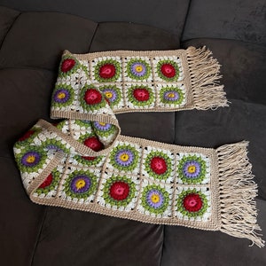 May include: A crocheted scarf with a floral pattern in shades of red, green, white, and purple. The scarf has a fringe on both ends.