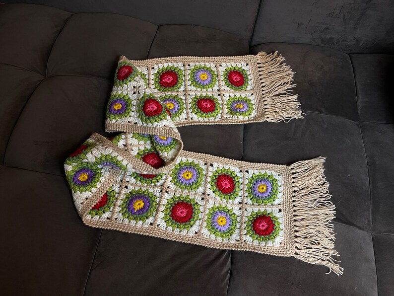 May include: A crocheted scarf with a floral pattern in shades of red, green, white, and purple. The scarf has a fringe on both ends.
