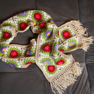 May include: A crocheted scarf with a fringe, featuring a floral pattern in shades of red, green, white, and purple.