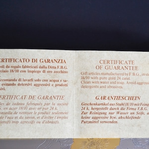 May include: A warranty certificate for gift articles made of steel with 24 karat gold. The certificate is printed in Italian, French, English, and German. It recommends cleaning with water and soap and avoiding aggressive detergents and abrasives.