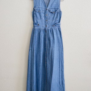 May include: A long, sleeveless denim dress with a button-up front and a V-neck. The dress has two pockets on the front and a tie at the waist.