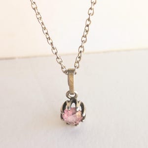 Op de afbeelding: Een zilverkleurige ketting met een kleine, ronde, roze edelsteen hanger. De hanger wordt op zijn plaats gehouden door een decoratieve zetting. De ketting is delicaat en hangt tegen een witte achtergrond. Een eenvoudig en elegant sieraad.