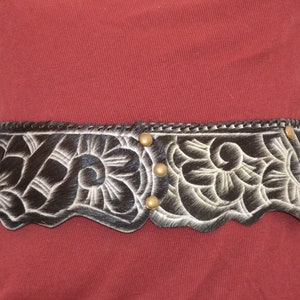 May include: A brown leather belt with a white floral design and gold-toned metal accents. The belt has a scalloped edge.