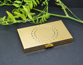 Puede incluir: Un estuche compacto de metal dorado con un diseño circular decorativo. El estuche tiene una tapa con bisagras y un pequeño cierre.