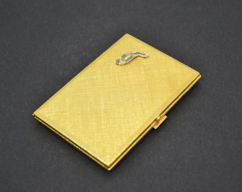 Puede incluir: Un estuche rectangular de color dorado con una superficie texturizada y un pequeño acento decorativo plateado. El estuche está cerrado y tiene un pequeño cierre lateral. El fondo es de color gris oscuro.