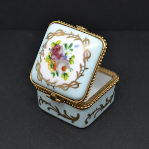 May include: A small, square, blue and gold trinket box with a floral design on the lid. The box is open and the inside is white.