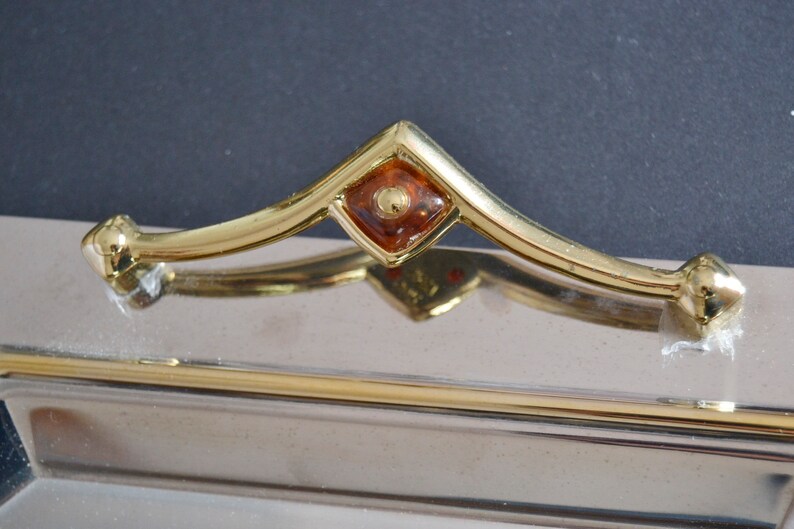 May include: A silver tray with a gold handle and a decorative amber-colored glass accent. The handle is curved and has a diamond-shaped design.