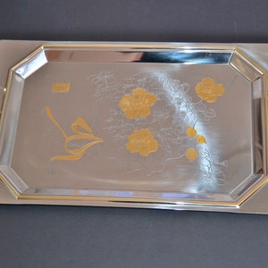 May include: A silver serving tray with gold accents and a floral design. The tray has two handles on the sides.
