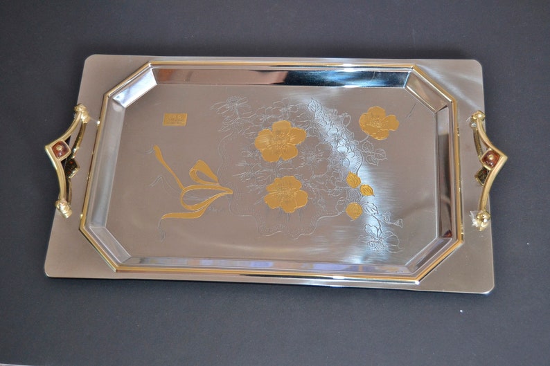 May include: A silver serving tray with gold accents and a floral design. The tray has two handles on the sides.