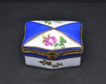 May include: A small, square porcelain trinket box with a blue and white geometric design. The lid features a floral pattern with a pink rose and green leaves. The box has gold accents around the edges and a gold hinge.