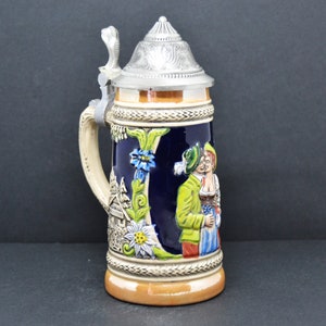 Puede incluir: Una jarra de cerveza de cerámica con un diseño floral azul y blanco. La jarra presenta una pareja con ropa tradicional alemana. La tapa es de peltre y tiene un diseño detallado.