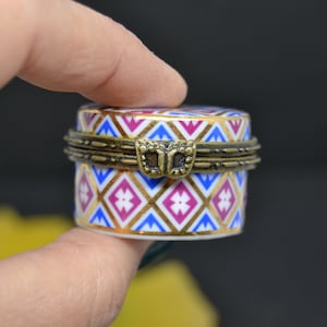 Puede incluir: Una pequeña caja redonda de porcelana con tapa con bisagras. La caja está decorada con un patrón geométrico en tonos azules, rosas, dorados y blancos. La tapa tiene una bisagra de latón y un cierre en forma de mariposa.